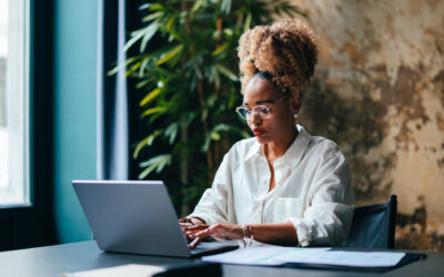 A young black female entrepreneur uses a laptop at a desk with papers spread out